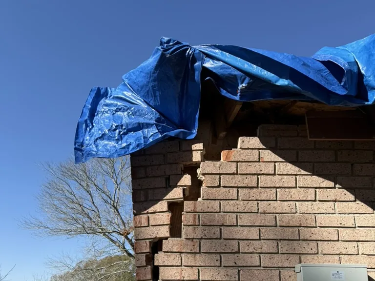 Brick house with a blue emergency tarp over a storm-broken roof corner and cracked brick below it